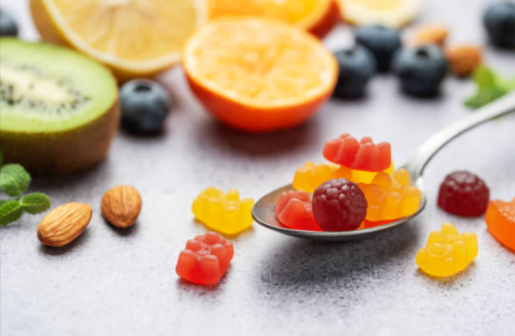 Colorful gummy vitamins on a spoon surrounded by fresh fruit slices like kiwi, orange, and blueberries